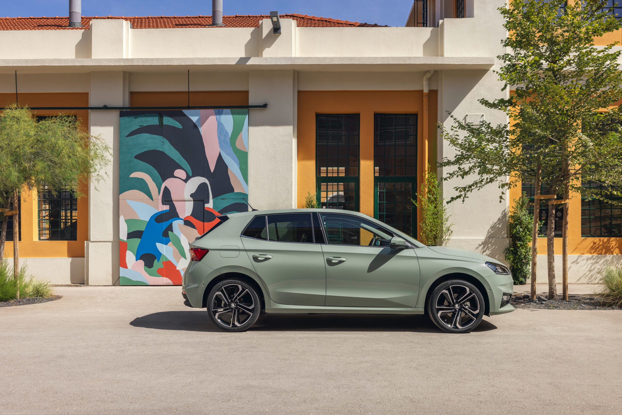 In diesem Bild ist ein Skoda Scala in hellgrün zu sehen. Das Fahrzueg steht vor einem Gebäude mit einem bunten Gemälde an der Hauswand. In dem Bild sind rechts und links Bäume gepflanzt.