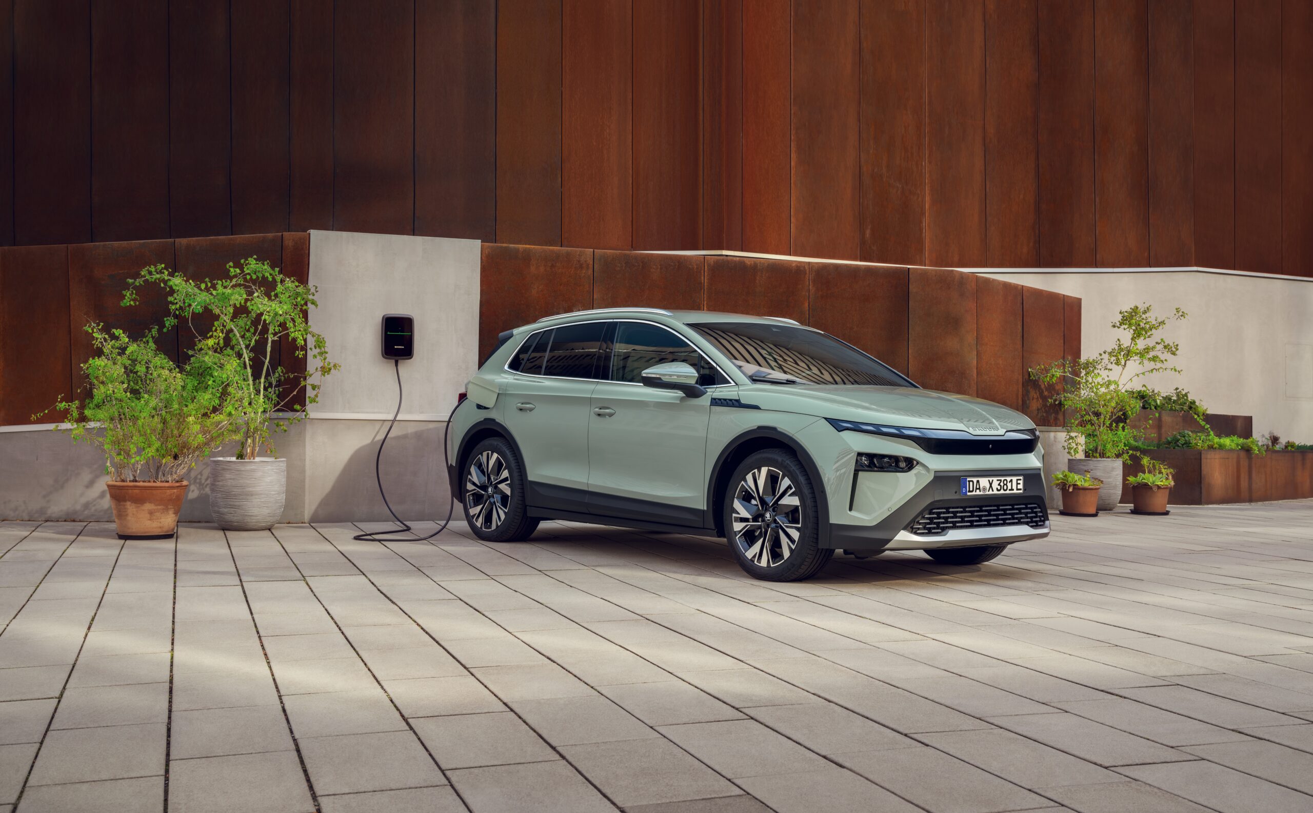 In dem Bild sieht man ein Elektrofahrzeug von Skoda. Das Fahrzeug ist an einer schwarzen Wallbox angeschlossen, die an einer Hauswand hängt. Das Haus ist in Rostoptik und vorne dran stehen grüne Pflanzen.