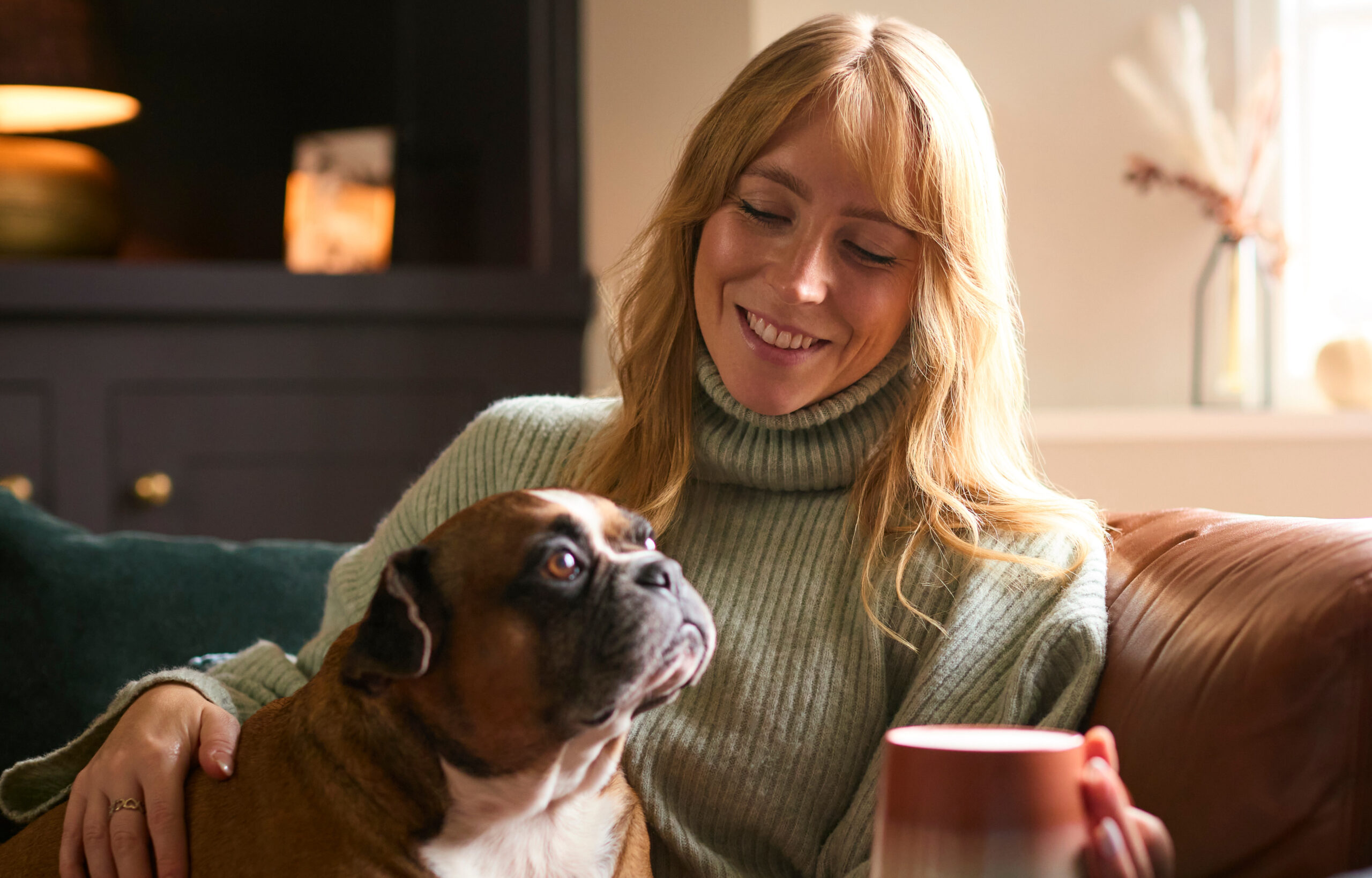 Eine Frau sitzt zu Hause auf dem Sofa. In der Hand hält sie eine Teetasse und auf dem Schoß hat sie einen Hund.