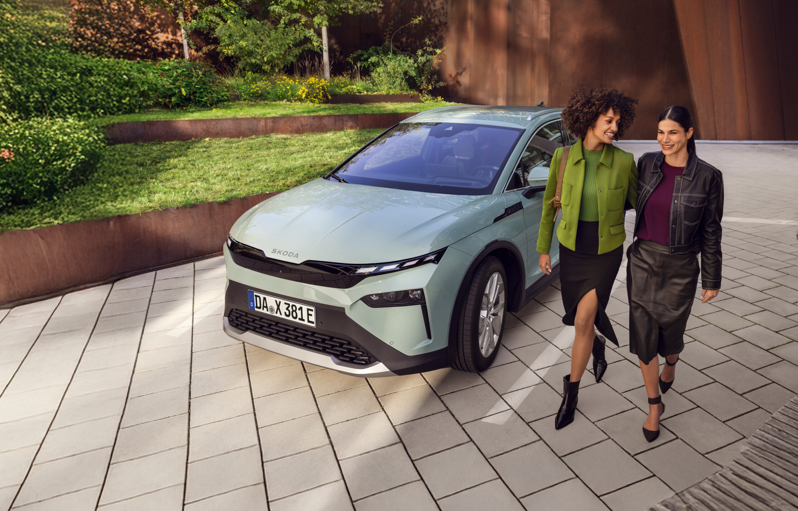 Zwei Frauen im Business-Outfit laufen Arm in Arm. Neben Ihnen ein Skoda Auto. Im Hintergrund eine Grünanlage.