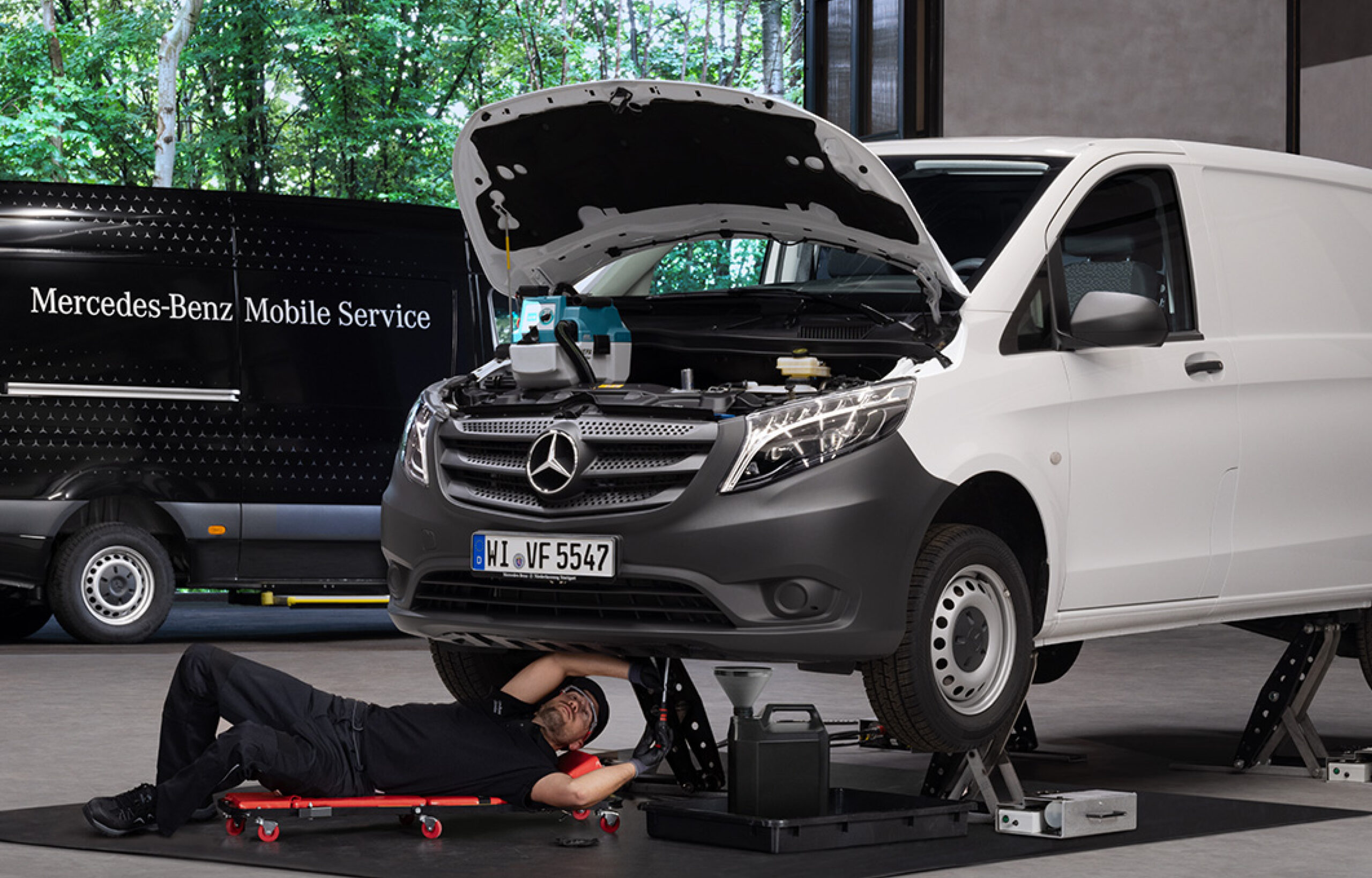 Auf dem Bild ist ein Van auf einer Hebebühne zu sehen un ddarunter liegt ein Monteur und schruabt am Unterboden des Fahrzeugs. Im Hintergrund is ein Sprinter mit der Aufschrift Mercedes-Benz Mobile Service zu sehen.