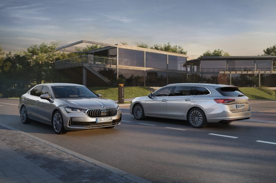 Auf dem Bild sind 2 Skoda Superb in silber zu sehen. Die Fahrzeuge fahren auf der Straße jewals in die andre richtung. Im Hintergrund ist ein Gebäude mit Glaswänden zu sehen.